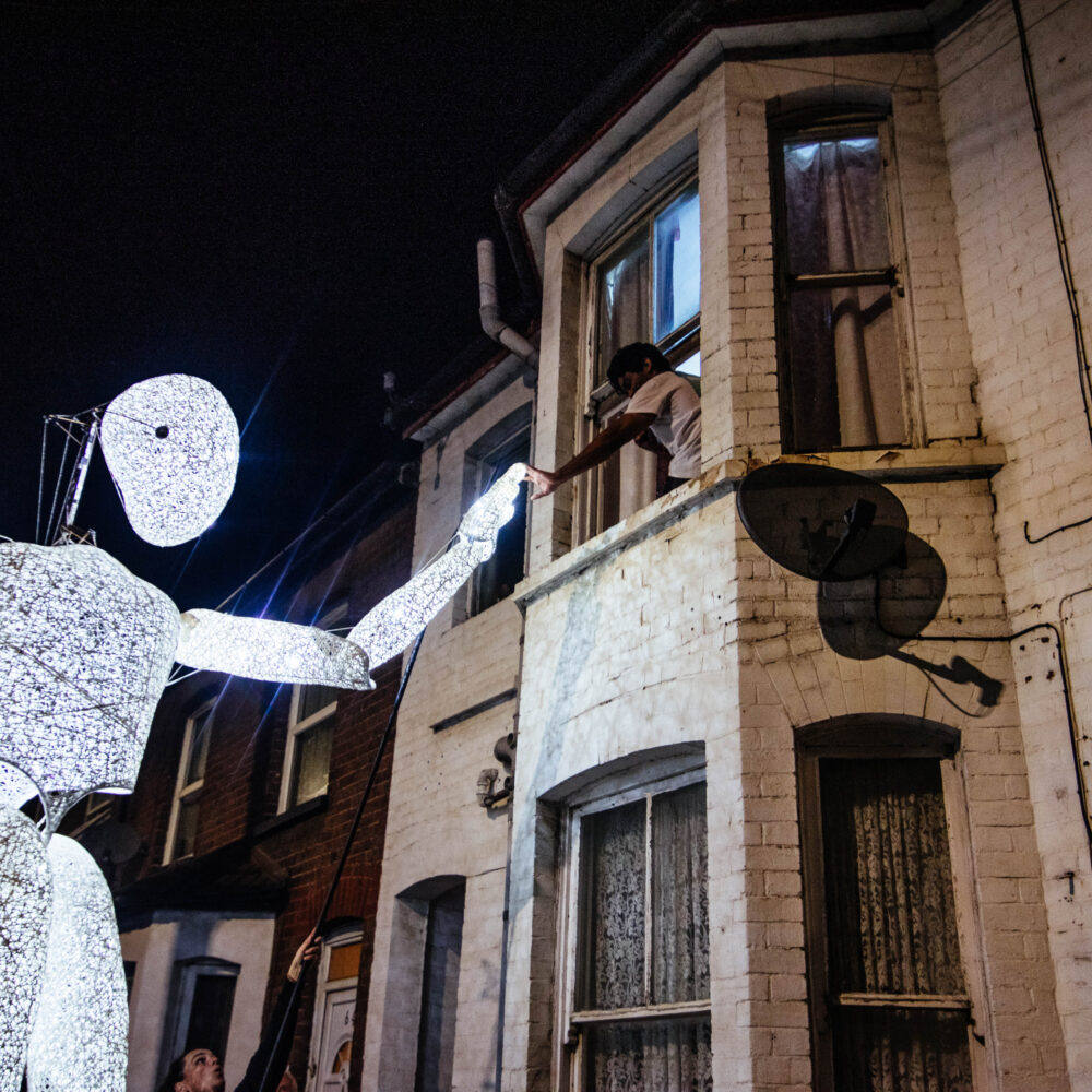 A giant illuminated figure high-fives a child through a house window