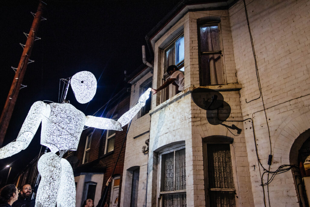 A giant illuminated figure high-fives a child through a house window