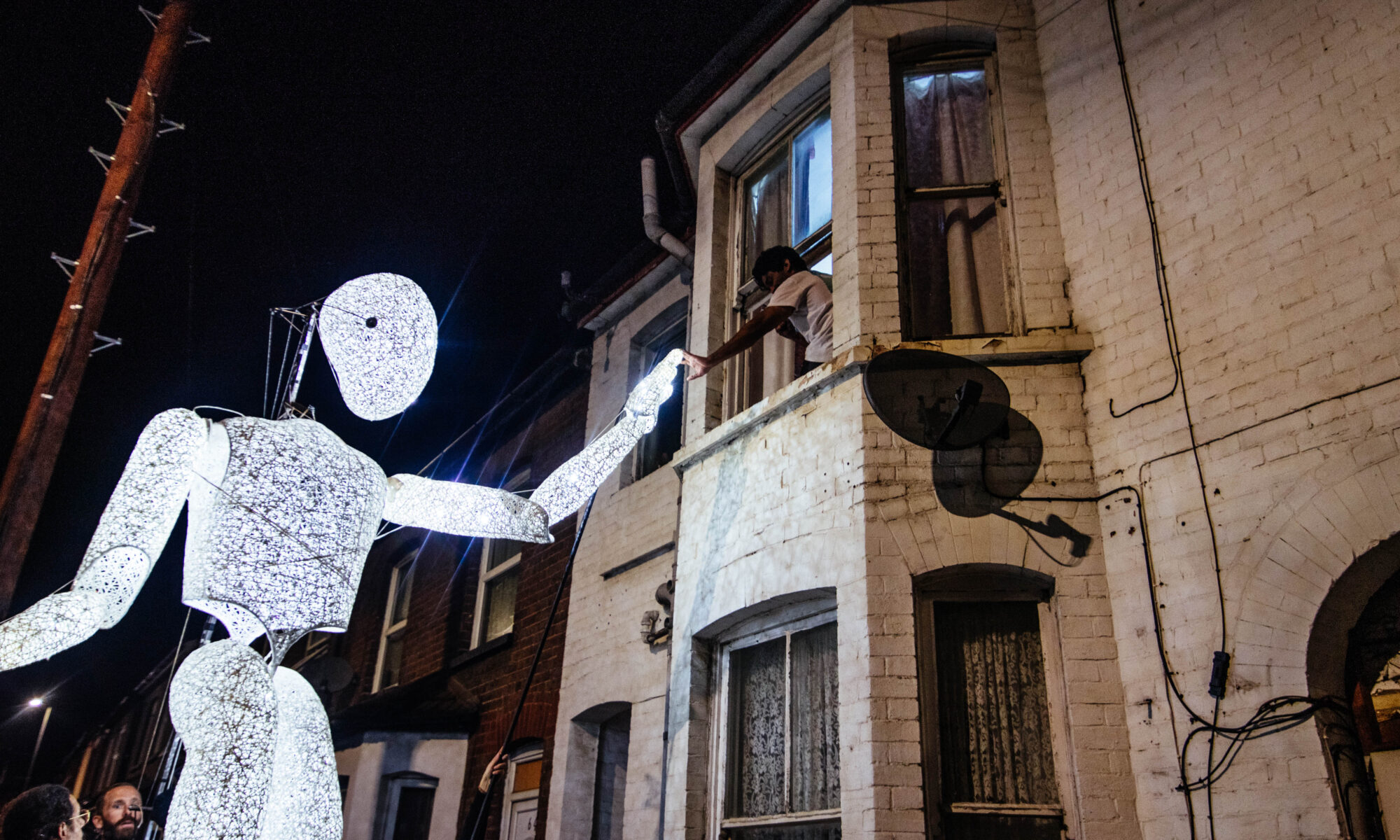 A giant illuminated figure high-fives a child through a house window