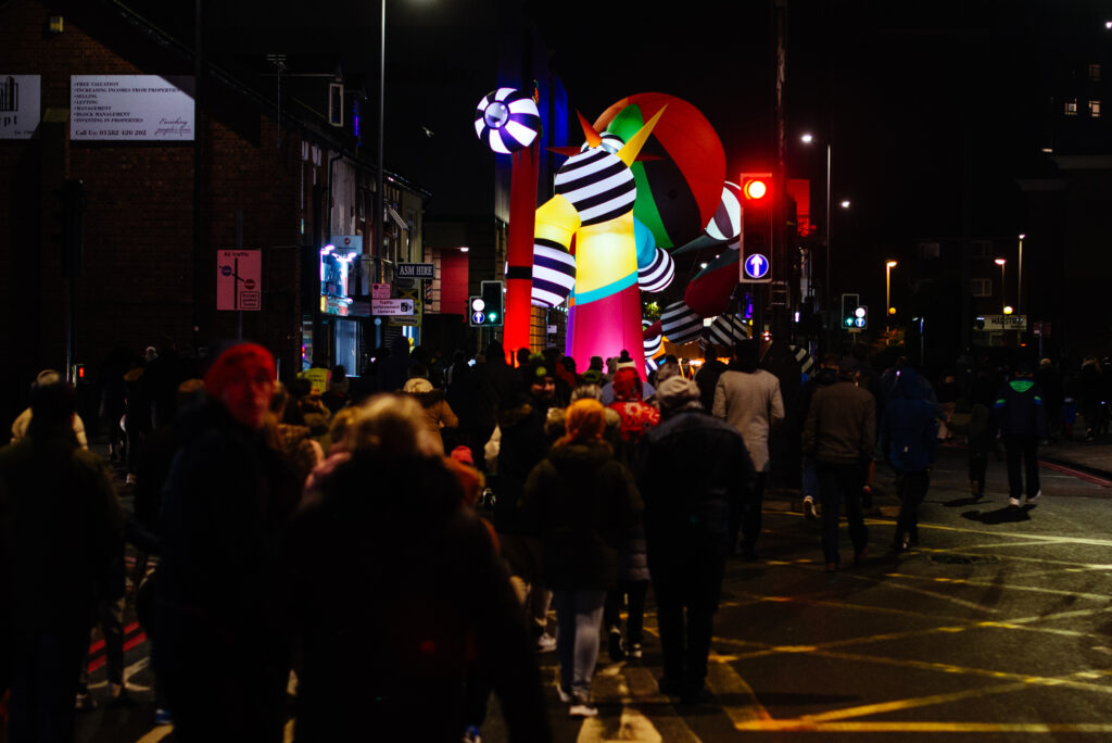A crowd follows an inflatable sculpture down a street at night