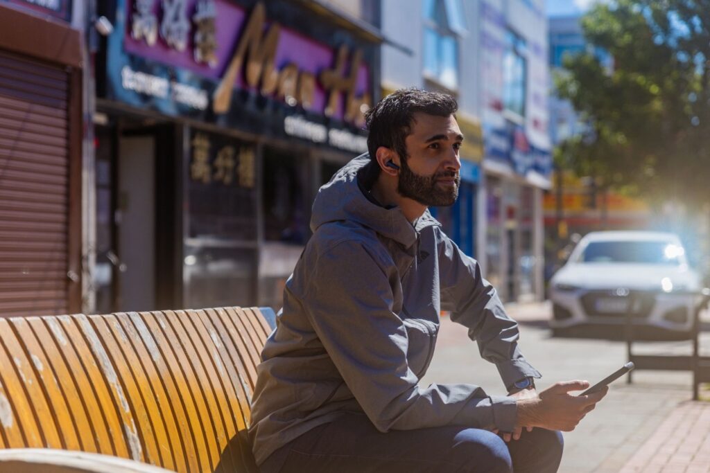 Man sitting on a bench on a high street listening to headphones